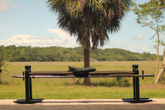 A 10' Charleston green joggling board with a black cushion.  Folded on top of the joggling board is a cover to protect the joggling board from the outside elements.  Behind the joggling board is overlooking the marsh on Edisto Island, SC.